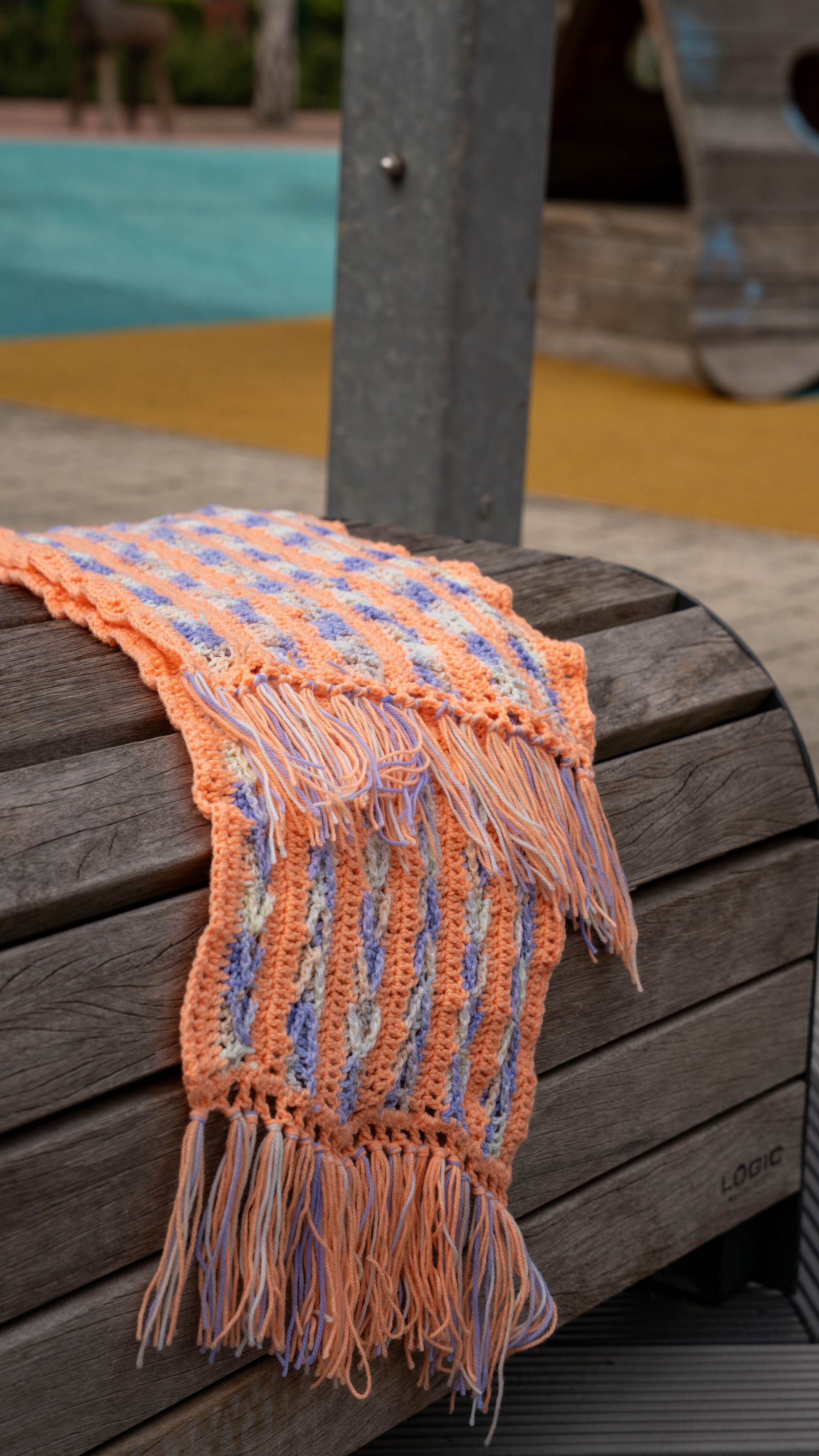 Peach, yellow and lilac crochet scarf laid flat on wooden bench showing fringe detail