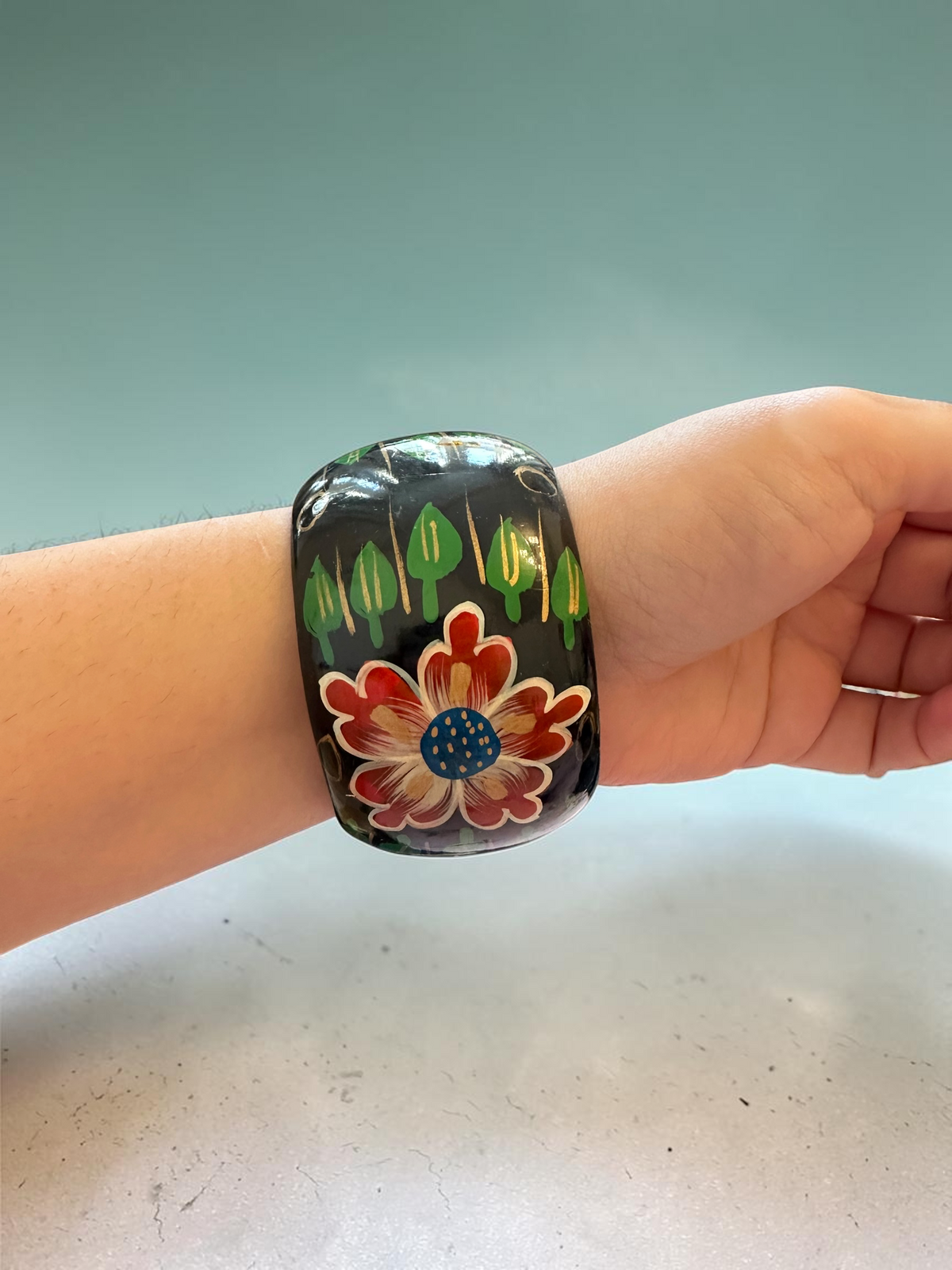 Hand-painted floral bangle worn on wrist, showing red flower with blue centre and green leaf motifs on black lacquer